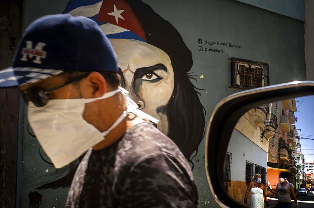 A man wearing a mask walks alongside a mural of Ernesto "Che" Guevara as other pedest ...