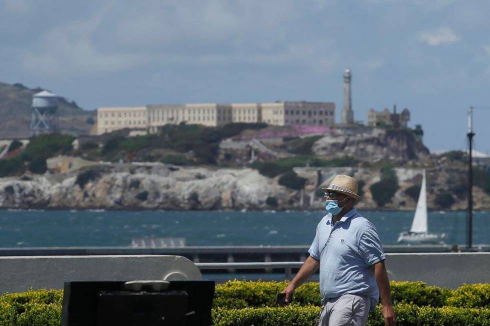 A man covers his face with a mask while walking on a path at Aquatic Park in front of Alcatraz ...