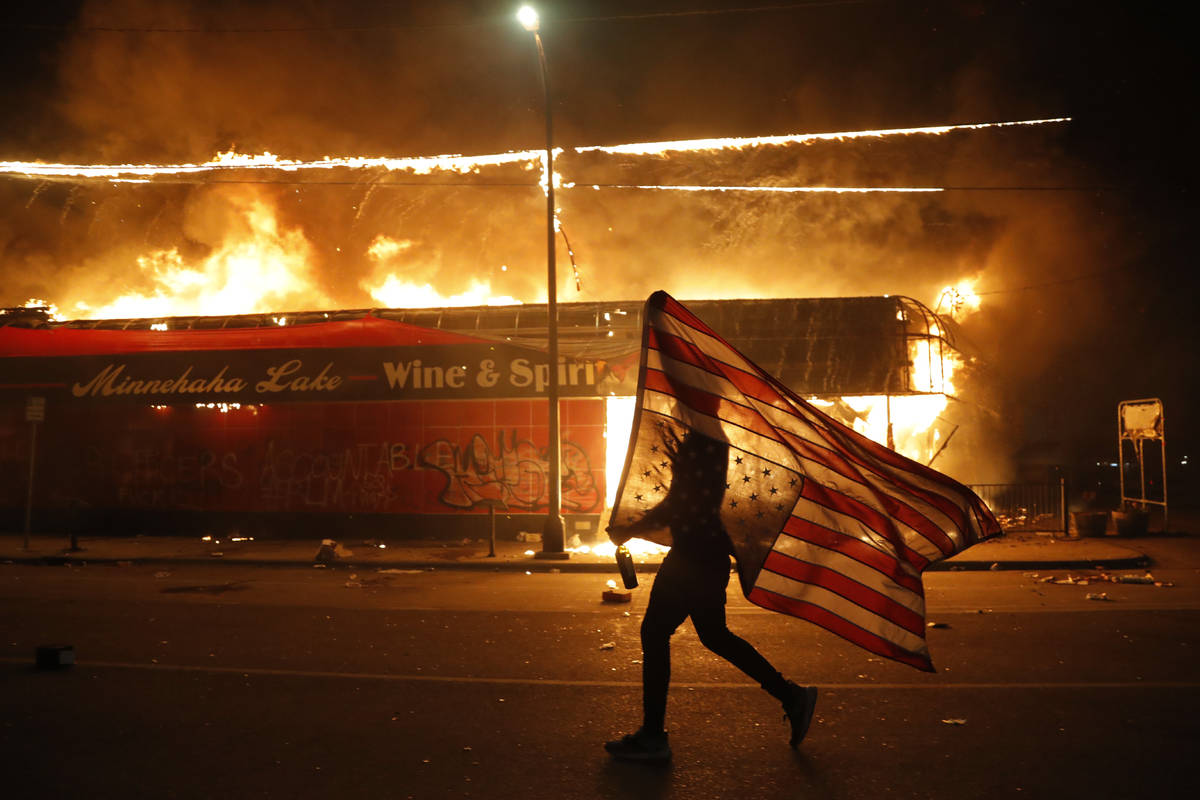 A protester carries the carries a U.S. flag upside, a sign of distress, next to a burning build ...