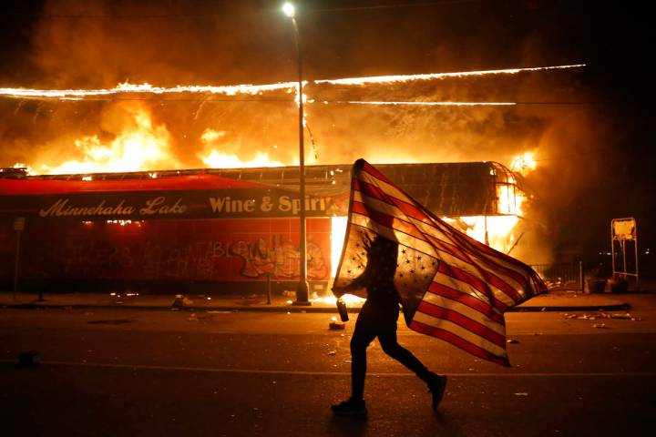 A protester carries the carries a U.S. flag upside, a sign of distress, next to a burning build ...
