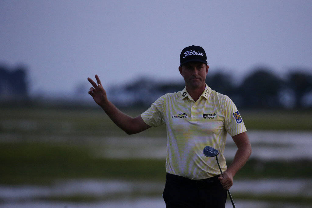 Webb Simpson waves as the sun disappears on the 18th green, on a course with no fans due to the ...