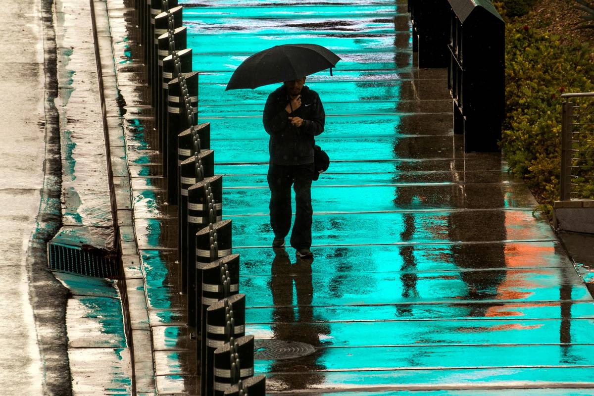A pedestrian is bathed in blue light from a lighted sign above as he walks in the rain about th ...