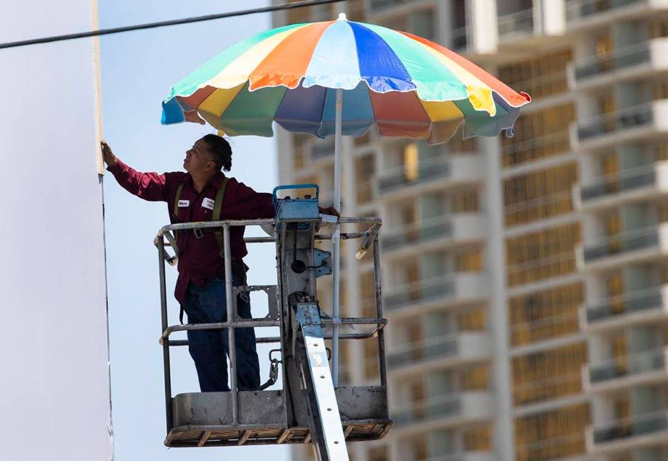 A worker uses a giant umbrella to protect himself from sun as he works on an outdoor advertisin ...