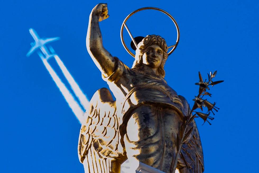 A passenger plane flies in clear blue sky and leaves a white trail as it flies over a statue in ...