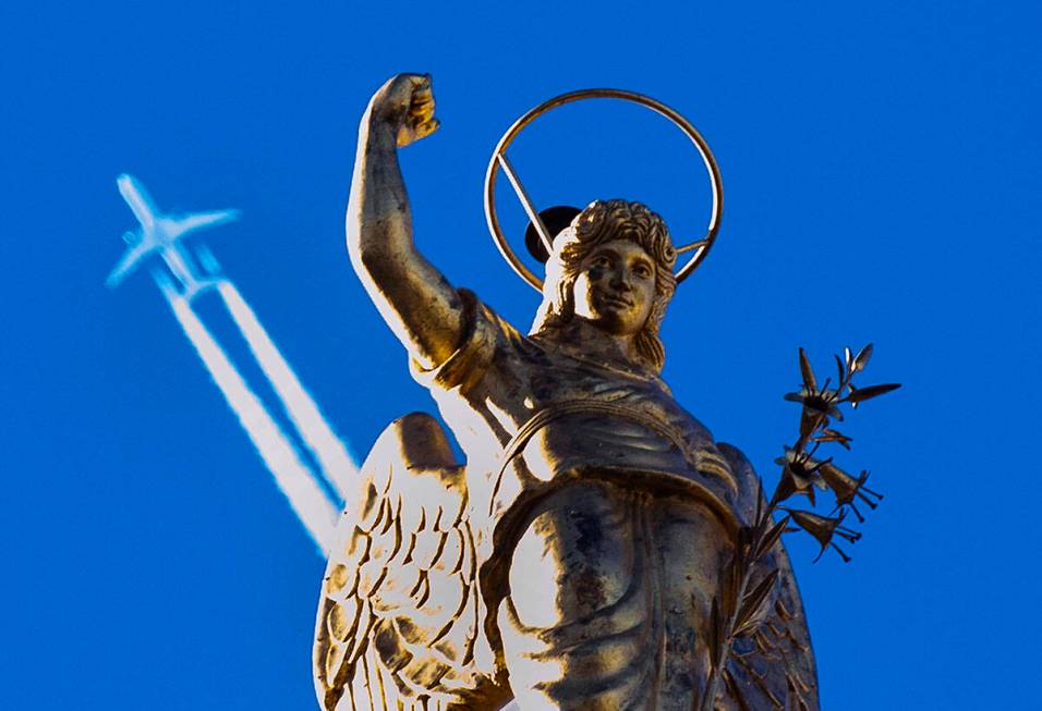 A passenger plane flies in clear blue sky and leaves a white trail as it flies over a statue at ...