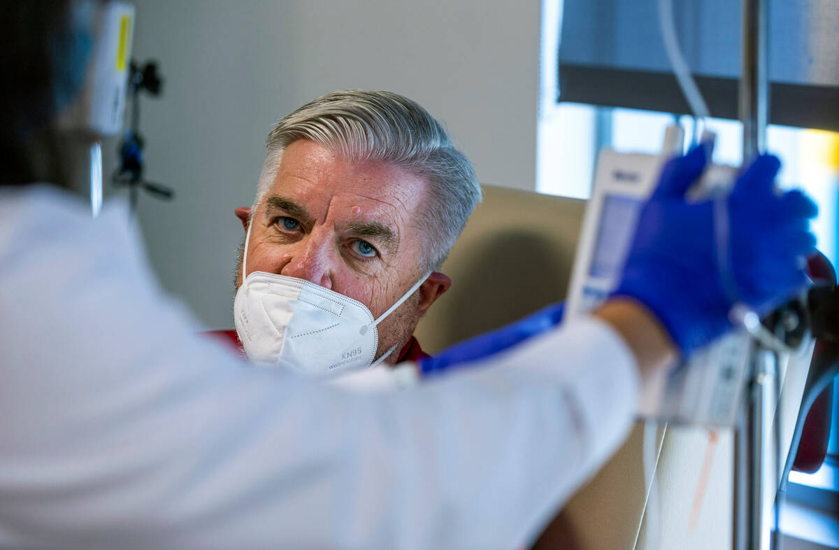 Clinical trial participant Bob Lathrop looks on as infusion room RN Betty Romero sets up his ne ...