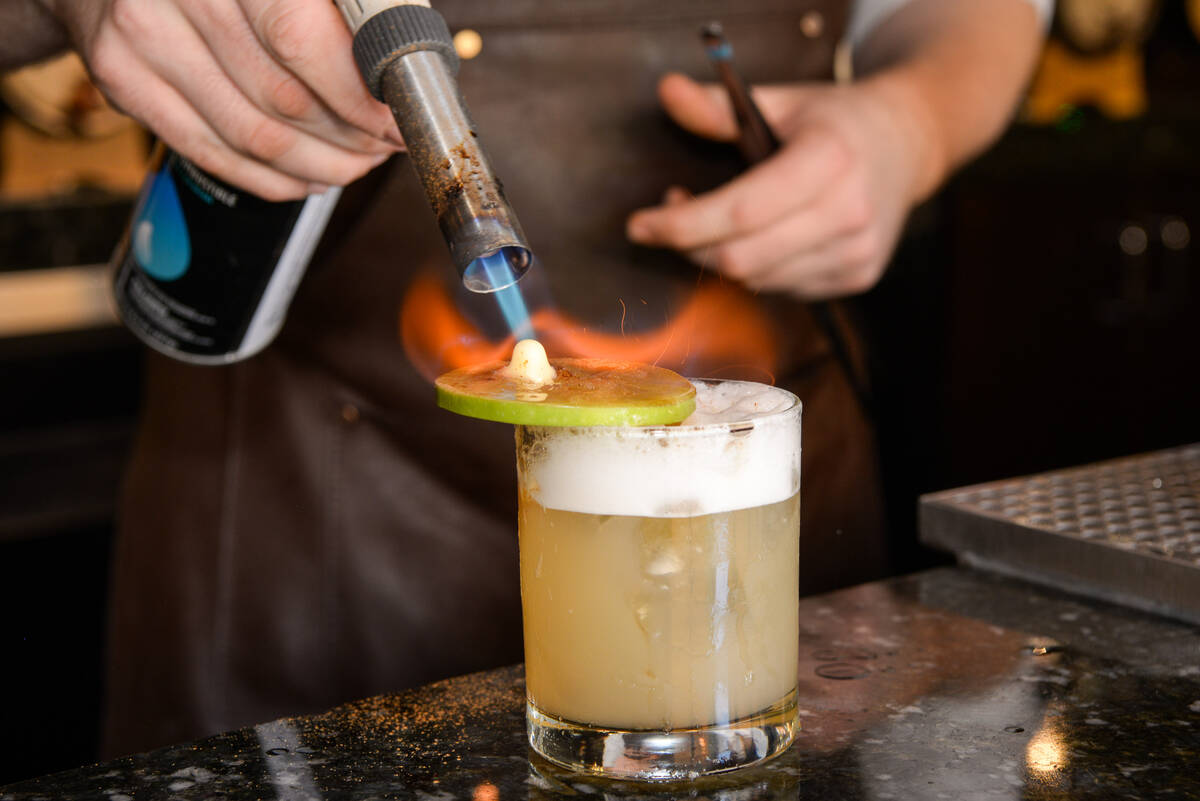 An apple slices is flamed with a kitchen torch to make the Apple Pie Harvest cocktail at Oak & ...