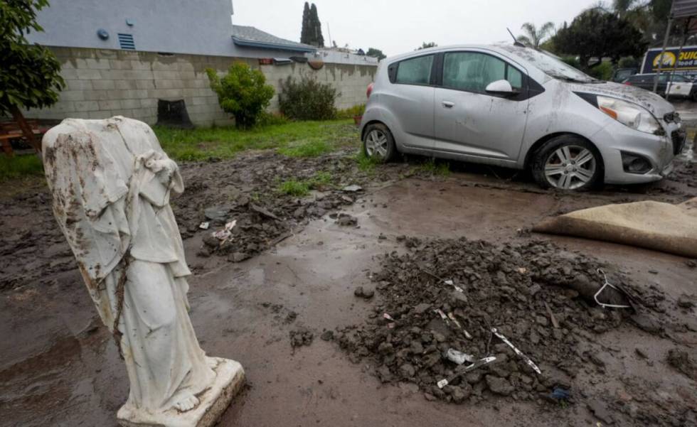 A broken statue and car covered in mud sit in front of a house damaged by a previous rainstorm ...