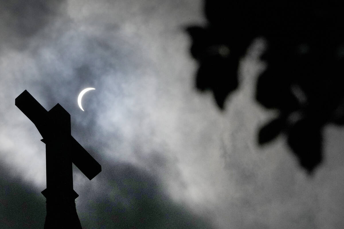 Clouds part as a partial eclipse of the sun and moon is seen atop the cross on the steeple of t ...