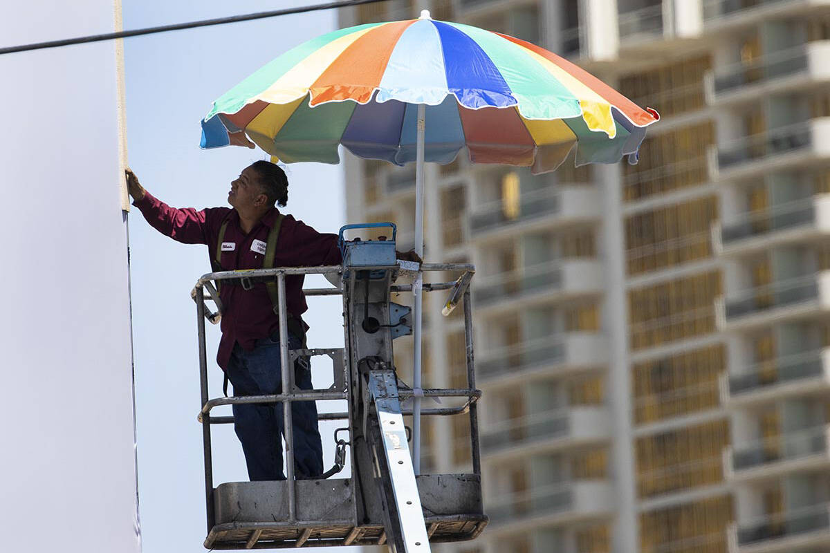 A worker uses a giant umbrella to protect himself from sun as he works on an outdoor advertisin ...