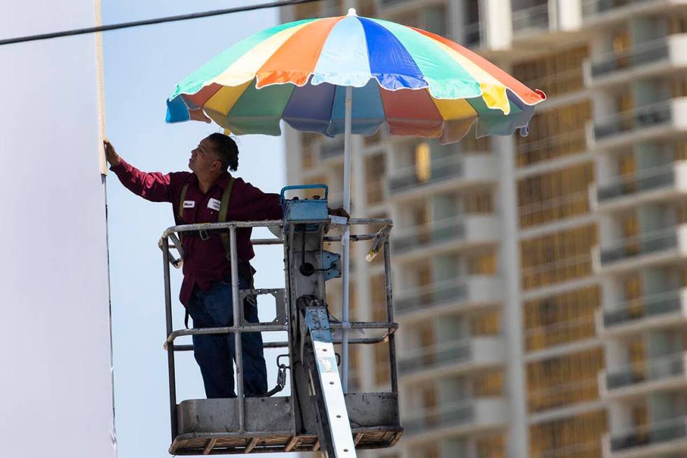 A worker uses a giant umbrella to protect himself from sun as he works on an outdoor advertisin ...