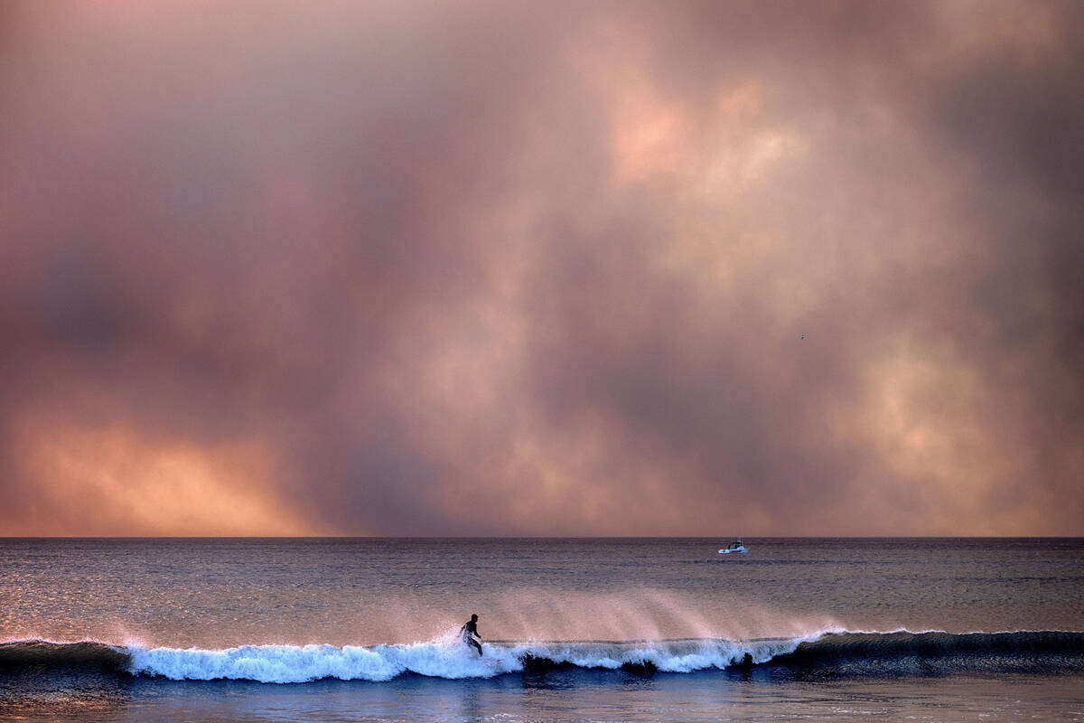 A surfer takes off on a wave under a blackened sky from the Palisades Fire in the Pacific Palis ...