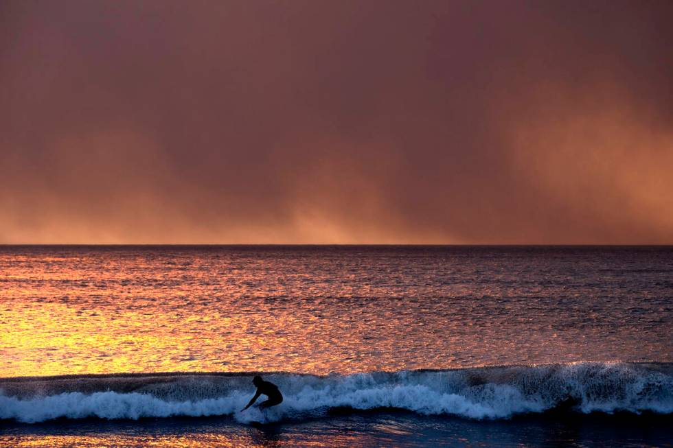 A surfer takes off on a wave under a blackened sky from the Palisades Fire in the Pacific Palis ...